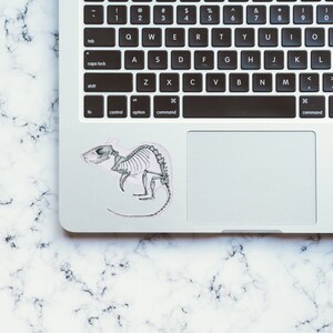 May include: A silver laptop computer with a black keyboard and a trackpad. A sticker of a rat skeleton is on the trackpad.