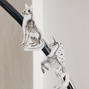 May include: Two skeletal animal figures, a dog and a unicorn, are attached to a silver metal rod. The dog is sitting and the unicorn is standing on its hind legs. Both figures are white with black outlines.