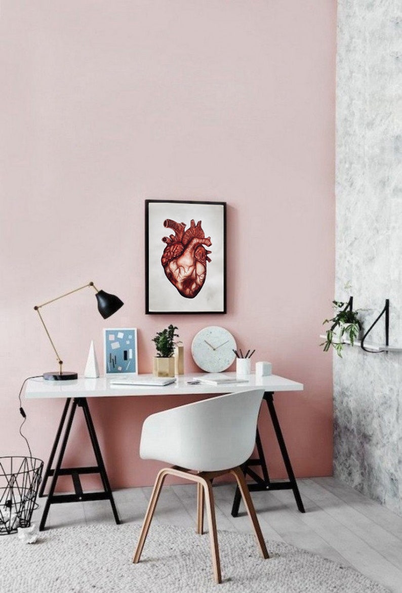 Puede incluir: Un escritorio blanco con patas de caballete negras, una silla blanca, una l&aacute;mpara dorada, un reloj y una impresi&oacute;n enmarcada de un coraz&oacute;n anat&oacute;mico rojo en una pared rosa.