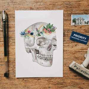 May include: A watercolor painting of a human skull with a floral crown. The skull is white and gray, and the flowers are pink, purple, yellow, and blue. The background is white.