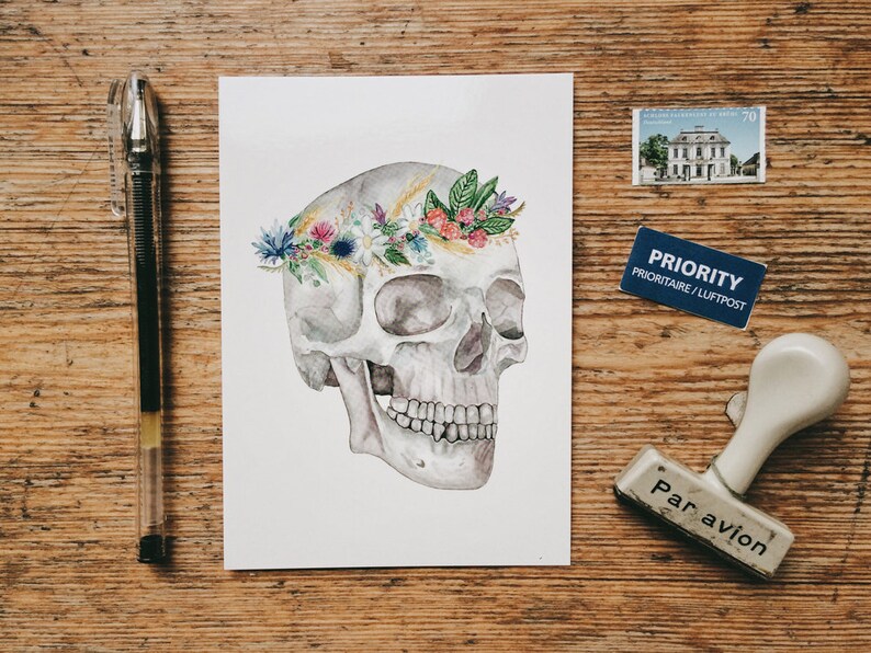 May include: A watercolor painting of a human skull with a floral crown. The skull is white and gray, and the flowers are pink, purple, yellow, and blue. The background is white.