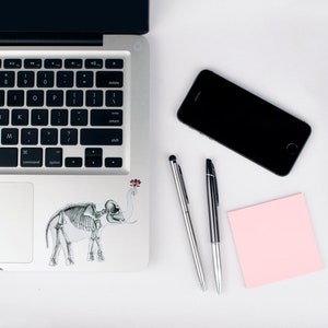 May include: A silver laptop computer with a black keyboard and a skeleton elephant sticker on the lid. There is a black smartphone, a pink sticky note, and two silver pens on a white surface.