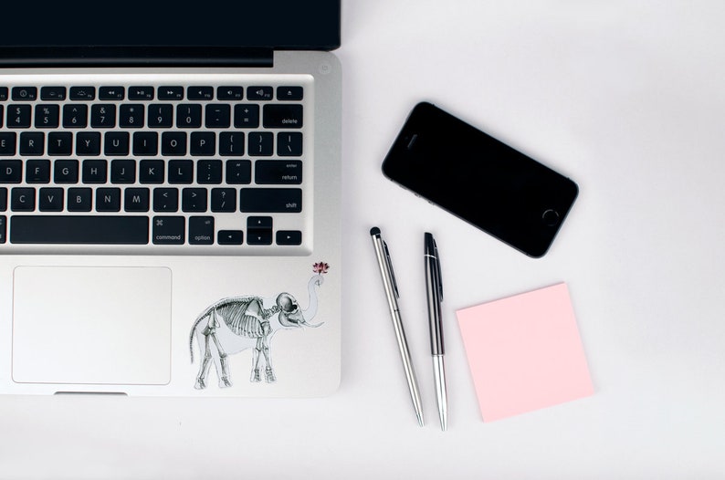 May include: A silver laptop computer with a black keyboard and a skeleton elephant sticker on the lid. There is a black smartphone, a pink sticky note, and two silver pens on a white surface.