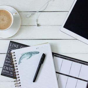 Pode incluir: Uma mesa branca com uma x&iacute;cara de caf&eacute;, um caderno com uma ilustra&ccedil;&atilde;o de uma baleia, uma caneta, um planejador preto e um tablet branco com uma tela preta.