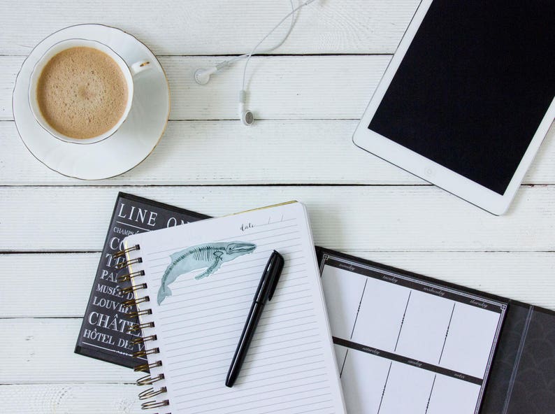 Pode incluir: Uma mesa branca com uma x&iacute;cara de caf&eacute;, um caderno com uma ilustra&ccedil;&atilde;o de uma baleia, uma caneta, um planejador preto e um tablet branco com uma tela preta.