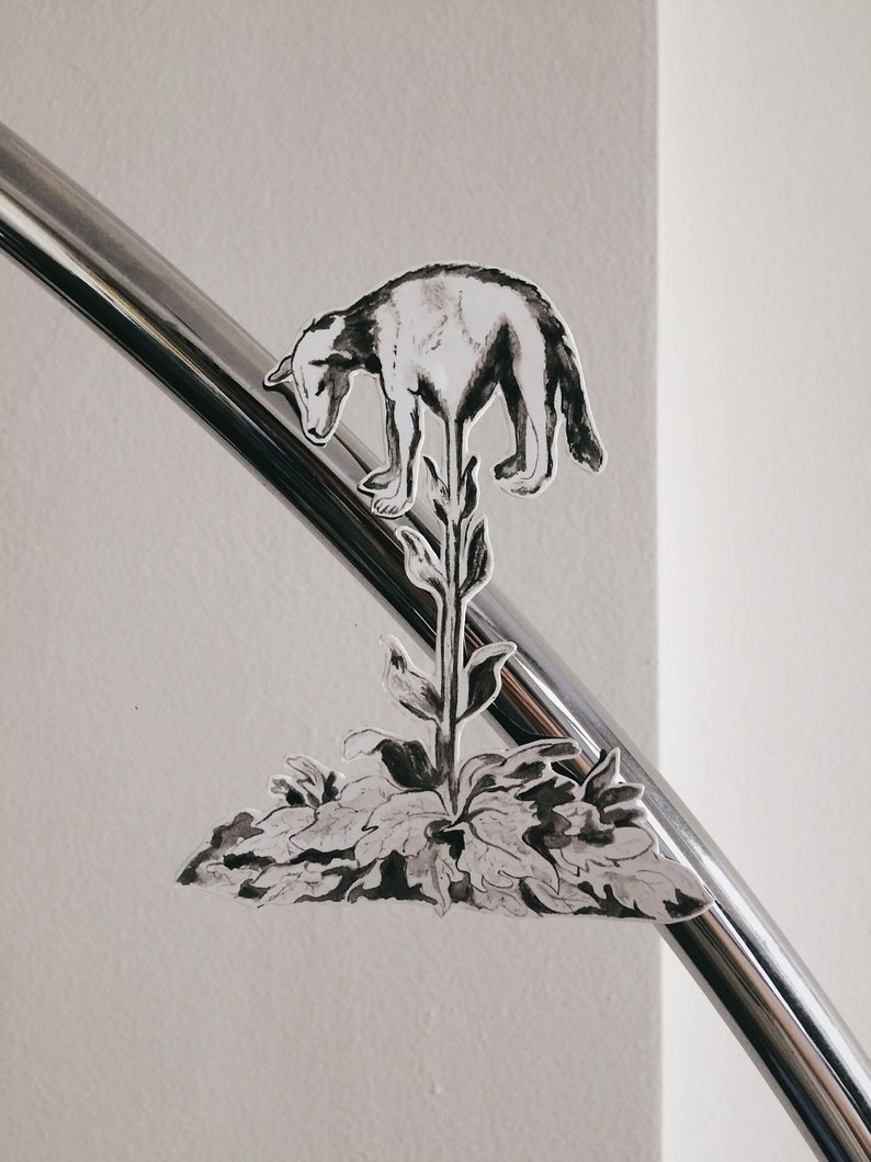 K&ouml;nnte beinhalten: Schwarzwei&szlig;-Illustration eines Hundes, der auf einer Pflanze mit Bl&auml;ttern steht. Die Illustration befindet sich auf einer silbernen Metalloberfl&auml;che.