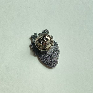 May include: A silver-colored pin shaped like a human heart, viewed from the back. The pin has a textured, dark gray surface and a silver clasp. The heart shape is detailed, with visible anatomical features.