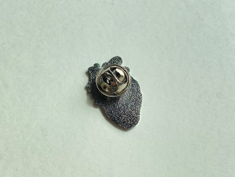 May include: A silver-colored pin shaped like a human heart, viewed from the back. The pin has a textured, dark gray surface and a silver clasp. The heart shape is detailed, with visible anatomical features.