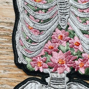 May include: Embroidered patch featuring a detailed skeleton design filled with pink and orange flowers. The patch has a black border and is made with gray, pink, orange, and green thread. The design is intricate and detailed.