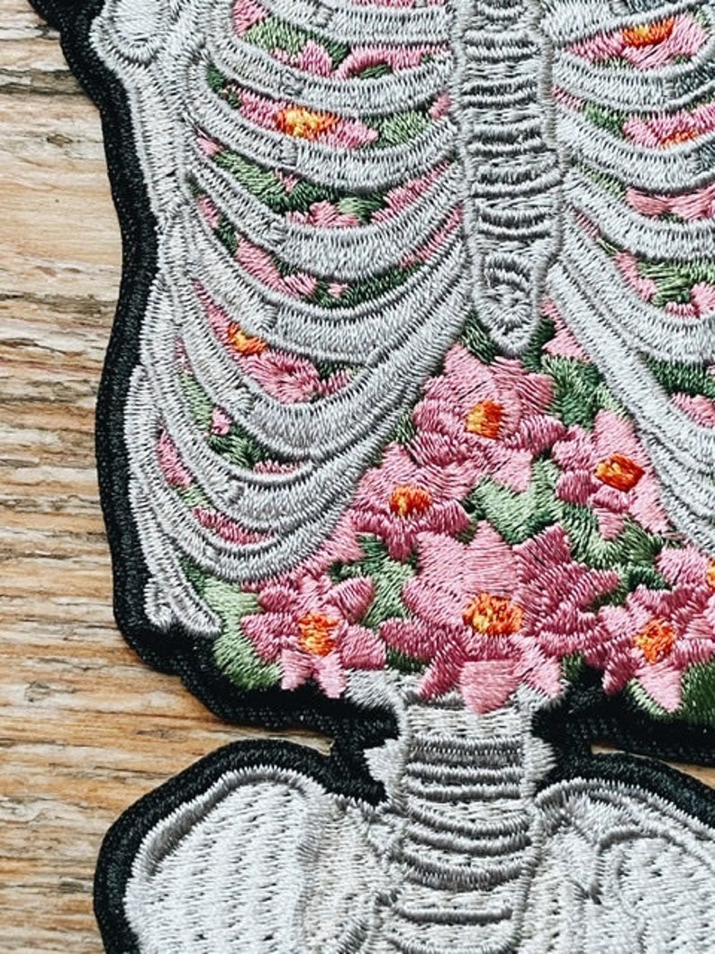 May include: Embroidered patch featuring a detailed skeleton design filled with pink and orange flowers. The patch has a black border and is made with gray, pink, orange, and green thread. The design is intricate and detailed.