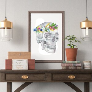 Pu&ograve; includere: Un dipinto ad acquerello di un teschio umano con una corona di fiori. Il teschio &egrave; bianco e i fiori sono colorati. Il dipinto &egrave; incorniciato e appeso a una parete sopra un tavolo di legno.