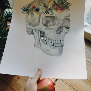 May include: Watercolour painting of a human skull wearing a floral crown made of wildflowers, wheat, and berries. The skull is painted in shades of grey and white, while the flowers are in shades of pink, purple, blue, and yellow.