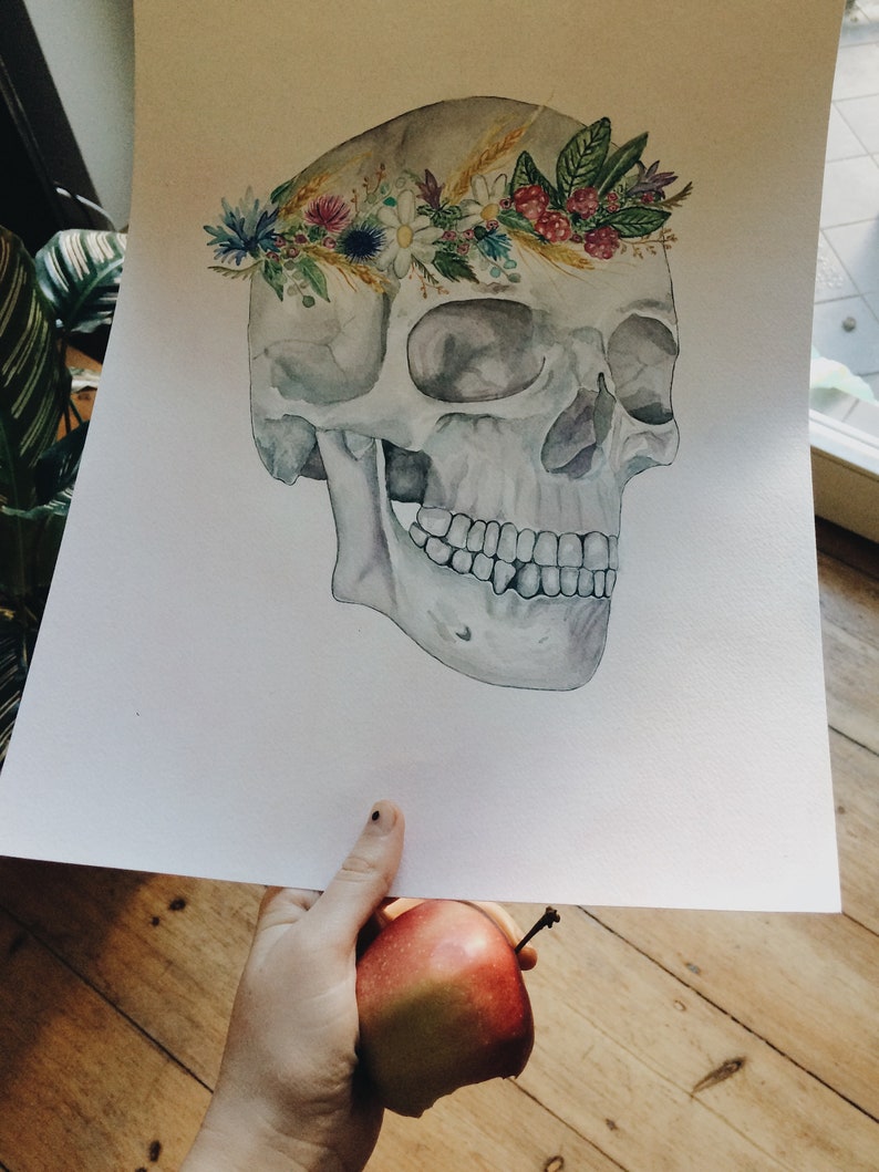 May include: Watercolour painting of a human skull wearing a floral crown made of wildflowers, wheat, and berries. The skull is painted in shades of grey and white, while the flowers are in shades of pink, purple, blue, and yellow.