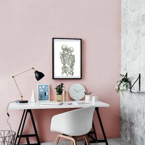 May include: A black framed print of two skeletons embracing and kissing, hanging on a light pink wall above a white desk with a white chair. The desk has a black lamp, a white vase with a plant, a clock, and a white pencil holder with pencils.