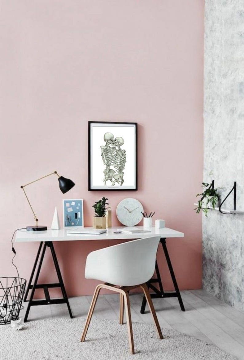 May include: A black framed print of two skeletons embracing and kissing, hanging on a light pink wall above a white desk with a white chair. The desk has a black lamp, a white vase with a plant, a clock, and a white pencil holder with pencils.