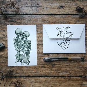 May include: Two skeletons are embracing and kissing. The skeletons are drawn in a green and white watercolor style. The image is on a white card. A white envelope with a black anatomical heart drawing is next to the card. A pen and a stamp are also on the wooden surface.