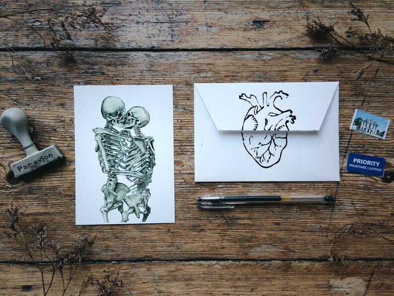 May include: Two skeletons are embracing and kissing. The skeletons are drawn in a green and white watercolor style. The image is on a white card. A white envelope with a black anatomical heart drawing is next to the card. A pen and a stamp are also on the wooden surface.