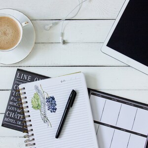 K&ouml;nnte beinhalten: Draufsicht auf einen Arbeitsplatz mit einer Kaffeetasse, einem Tablet und Notizb&uuml;chern. Eine wei&szlig;e Kaffeetasse mit Goldrand steht auf einer Untertasse. Ein schwarzer Stift liegt auf einem Spiralnotizbuch mit einer Traubenillustration. Wei&szlig;e Ohrh&ouml;rer sind ebenfalls vorhanden.