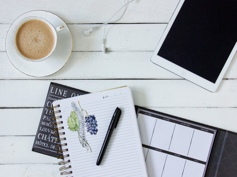 K&ouml;nnte beinhalten: Draufsicht auf einen Arbeitsplatz mit einer Kaffeetasse, einem Tablet und Notizb&uuml;chern. Eine wei&szlig;e Kaffeetasse mit Goldrand steht auf einer Untertasse. Ein schwarzer Stift liegt auf einem Spiralnotizbuch mit einer Traubenillustration. Wei&szlig;e Ohrh&ouml;rer sind ebenfalls vorhanden.