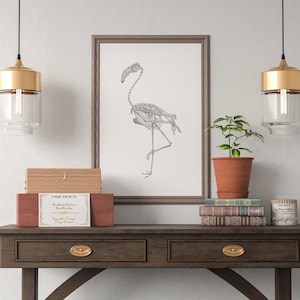 May include: A framed print of a flamingo skeleton drawing hangs above a wooden console table with two drawers. Two gold pendant lights with clear glass shades hang above the table. A small potted plant sits on the table next to a stack of books and a candle.
