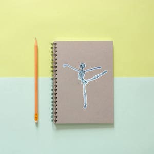 May include: A brown notebook with a spiral binding and a sticker of a skeleton dancing on the front. A yellow pencil is next to the notebook.