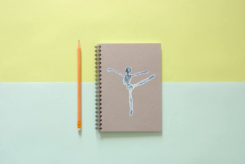 May include: A brown notebook with a spiral binding and a sticker of a skeleton dancing on the front. A yellow pencil is next to the notebook.