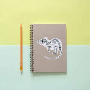 May include: A gray notebook with a spiral binding and a sticker of a rat skeleton on the front. A yellow pencil is lying next to the notebook.