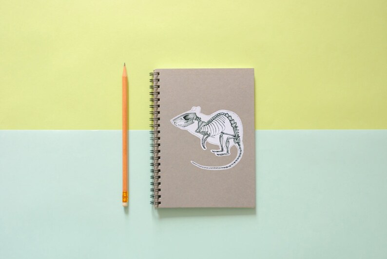 May include: A gray notebook with a spiral binding and a sticker of a rat skeleton on the front. A yellow pencil is lying next to the notebook.