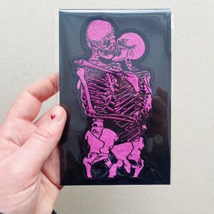 K&ouml;nnte beinhalten: Rosa gestickter Patch mit zwei Skeletten, die sich umarmen und k&uuml;ssen. Der Patch befindet sich auf einem schwarzen Hintergrund.