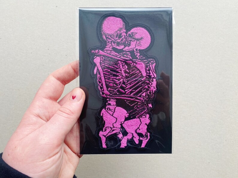 K&ouml;nnte beinhalten: Rosa gestickter Patch mit zwei Skeletten, die sich umarmen und k&uuml;ssen. Der Patch befindet sich auf einem schwarzen Hintergrund.