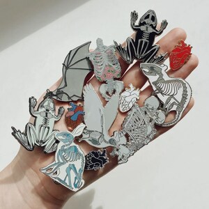 May include: A collection of enamel pins featuring anatomical designs. The pins depict skeletons of various animals, including a frog, bat, and rabbit, along with heart and ribcage designs. The pins are in shades of gray, white, black, red, and blue.