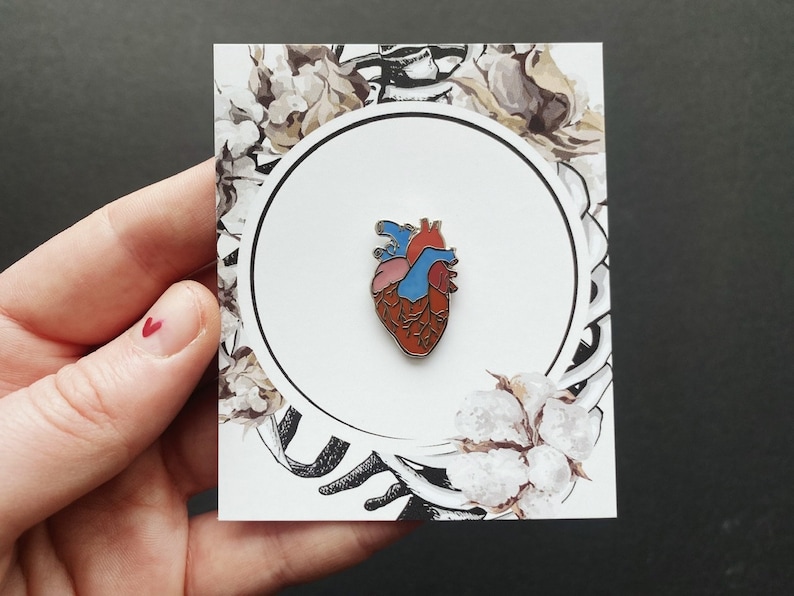 May include: A red, blue, and white anatomical heart enamel pin on a white card with a floral design.
