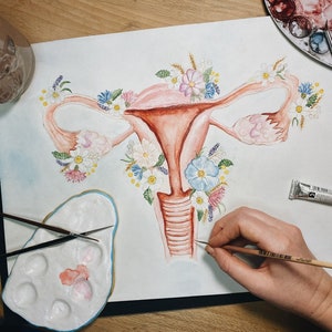 May include: A watercolor painting of a uterus with flowers surrounding it. The painting is on a white sheet of paper and is being worked on by an artist.