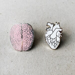K&ouml;nnte beinhalten: Zwei Emaille-Pins, einer zeigt ein rosa Gehirn und der andere ein wei&szlig;es Herz.