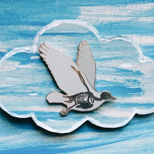 Puede incluir: Un pin de esmalte blanco de un p&aacute;jaro con una estructura esquel&eacute;tica visible en su interior. El pin tiene forma de nube y est&aacute; sobre un fondo azul.