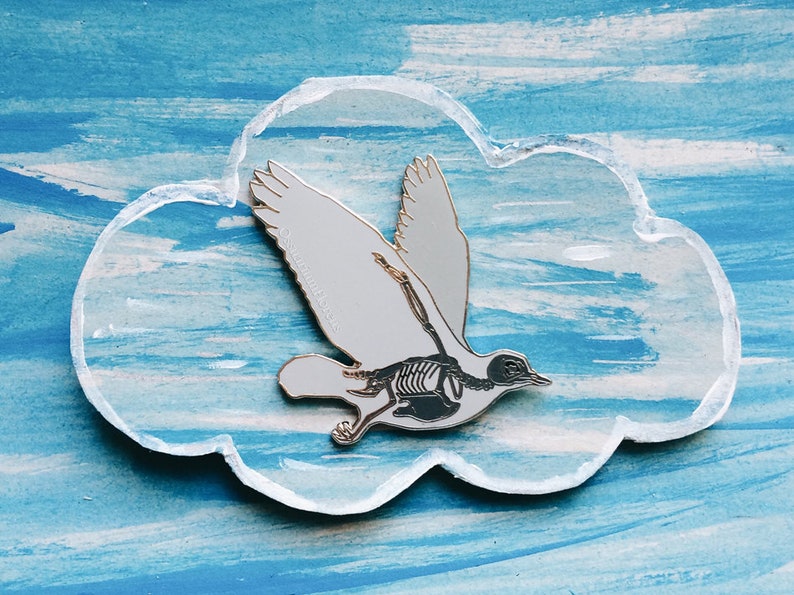 Puede incluir: Un pin de esmalte blanco de un p&aacute;jaro con una estructura esquel&eacute;tica visible en su interior. El pin tiene forma de nube y est&aacute; sobre un fondo azul.