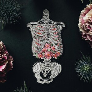 May include: Embroidered patch featuring a human rib cage filled with pink flowers. The patch is gray and white with black outlining. The background is black, with pink and purple flowers and green foliage surrounding the patch.