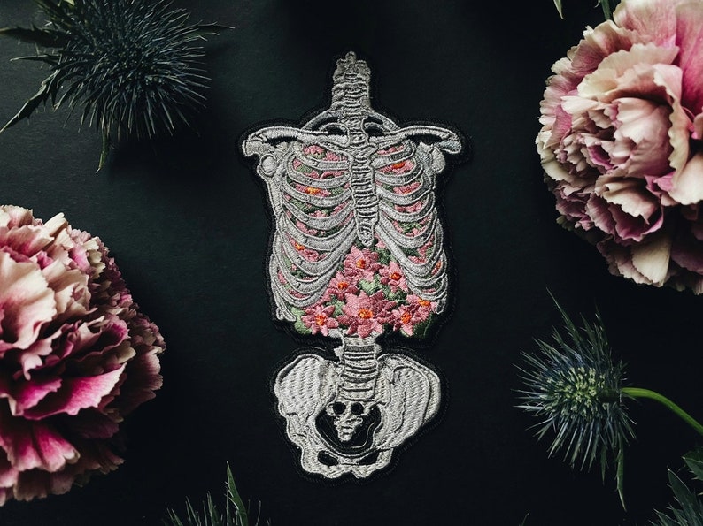 May include: Embroidered patch featuring a human rib cage filled with pink flowers. The patch is gray and white with black outlining. The background is black, with pink and purple flowers and green foliage surrounding the patch.