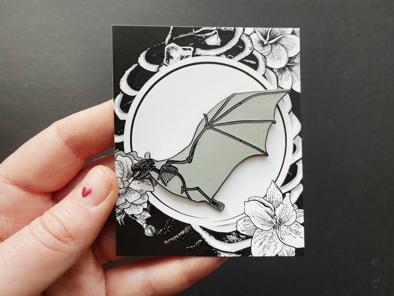 Puede incluir: Un pin de esmalte blanco y negro con un murci&eacute;lago estilizado con una envergadura de alas gris. El pin est&aacute; sobre un fondo negro con dise&ntilde;os florales blancos.