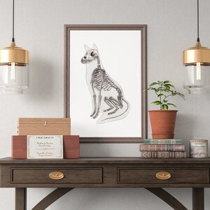 K&ouml;nnte beinhalten: Eine gerahmte Schwarzwei&szlig;illustration eines sitzenden Hundeskeletts auf wei&szlig;em Hintergrund. Der Rahmen ist braun und h&auml;ngt an einer wei&szlig;en Wand. Auf einem braunen Holztisch mit zwei Schubladen steht eine Holzkiste mit dem Etikett "DOG OFFICE" und eine kleine Topfpflanze.