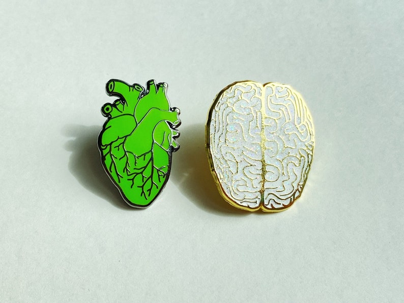 May include: Two enamel pins: a green anatomical heart with black outlines and a white brain with gold outlines. The pins are set against a light background. These pins are ideal for medical students or science enthusiasts.