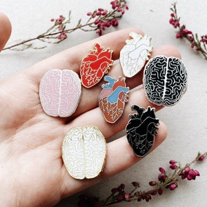 Puede incluir: Un conjunto de seis pines de esmalte que representan corazones y cerebros anat&oacute;micos. Los pines son de varios colores, incluyendo rojo, rosa, negro, blanco y azul. Los pines se muestran en una mano.