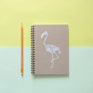 Peut inclure: Un carnet brun avec une reliure spirale et une illustration de squelette de flamant rose blanc sur la couverture. Un crayon jaune est pos&eacute; &agrave; c&ocirc;t&eacute; du carnet sur un fond vert clair et jaune.