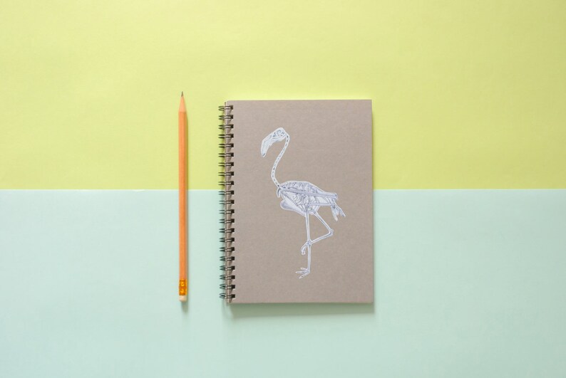 Peut inclure: Un carnet brun avec une reliure spirale et une illustration de squelette de flamant rose blanc sur la couverture. Un crayon jaune est pos&eacute; &agrave; c&ocirc;t&eacute; du carnet sur un fond vert clair et jaune.