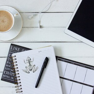 Op de afbeelding: Een witte houten tafel met een kop koffie, een notitieboek met een pen en een tekening van een skelet van een kikker, een zwart-witte planner en een witte tabletcomputer met een zwart scherm.
