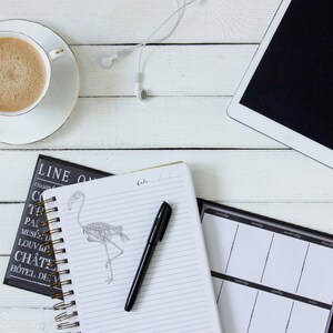 Peut inclure: Une table en bois blanc avec une tasse de caf&eacute;, un carnet avec un stylo, un agenda noir et blanc et une tablette blanche avec des &eacute;couteurs noirs.