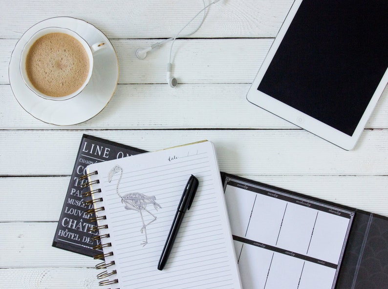 Peut inclure: Une table en bois blanc avec une tasse de caf&eacute;, un carnet avec un stylo, un agenda noir et blanc et une tablette blanche avec des &eacute;couteurs noirs.