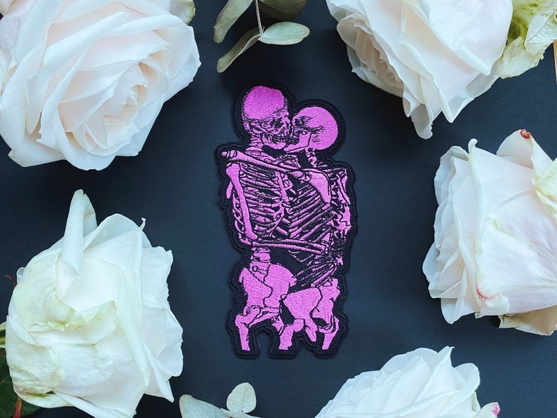 K&ouml;nnte beinhalten: Rosa gestickter Patch mit zwei sich umarmenden Skeletten vor schwarzem Hintergrund. Die Skelette befinden sich in einer romantischen Pose, wobei der Kopf des weiblichen Skeletts auf der Schulter des m&auml;nnlichen Skeletts ruht. Der Patch ist von wei&szlig;en Rosen umgeben.