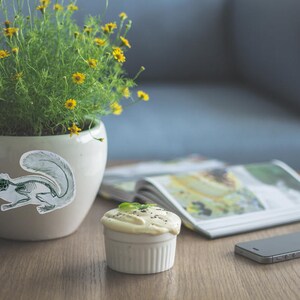 K&ouml;nnte beinhalten: Ein cremefarbener Keramiktopf mit einem Eichh&ouml;rnchen-Skelett-Aufkleber enth&auml;lt eine gr&uuml;ne Pflanze mit gelben Bl&uuml;ten. Ein kleiner wei&szlig;er Ramekin mit einem Dessert und ein Smartphone stehen auf einem Holztisch. Eine Zeitschrift ist im Hintergrund ge&ouml;ffnet.