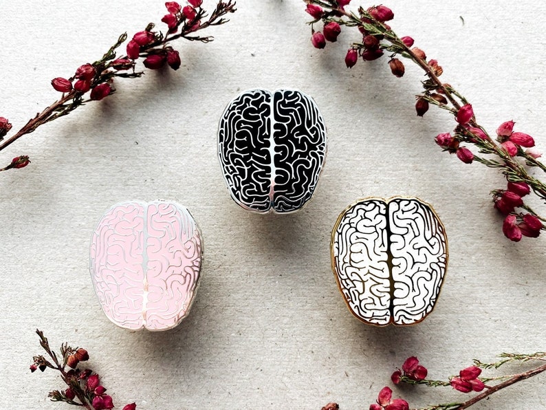 May include: Three enamel pins shaped like a human brain. One pin is black and white, one is pink and white, and one is gold and white.